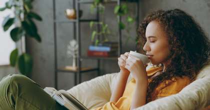 A woman with curly hair is reading a book and drinking from a cup.