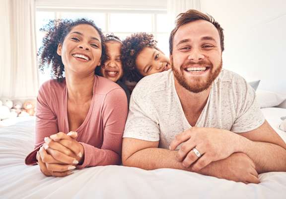 A family of three is lying on a bed, smiling and looking at the camera.