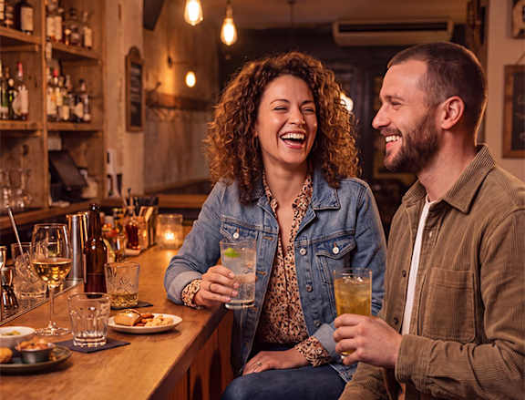 A man and a woman are sitting at a bar, laughing and enjoying drinks.
