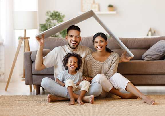 A family of three is sitting on the floor holding a paper house together.