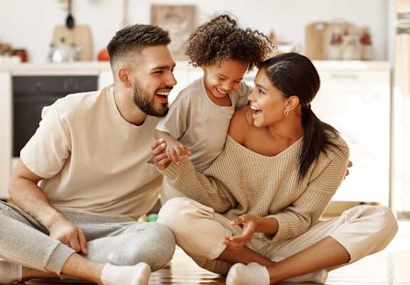 A man and a woman are sitting on the floor with a child between them, all are smiling and appear to be happy.