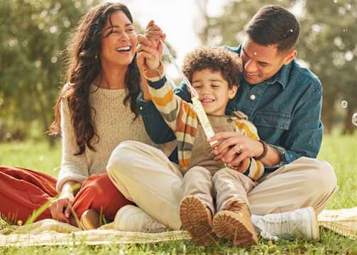 A family of three is sitting on a blanket in a park, blowing bubbles.