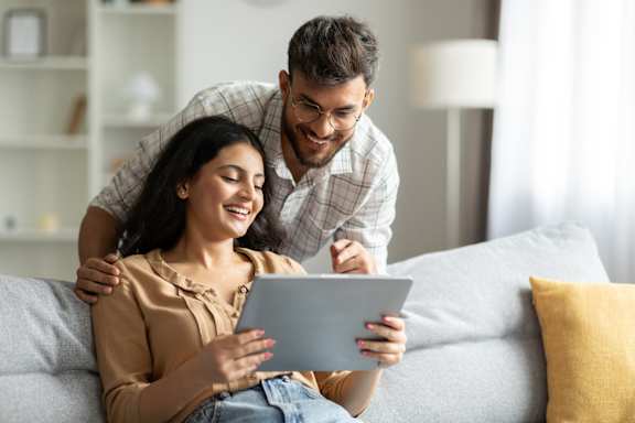 A man and a woman are sitting on a couch, with the woman holding a tablet and the man standing behind her.