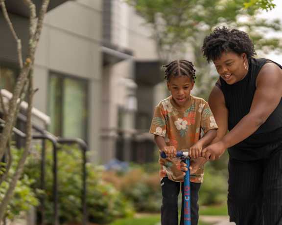 A woman is teaching a young girl how to ride a scooter.