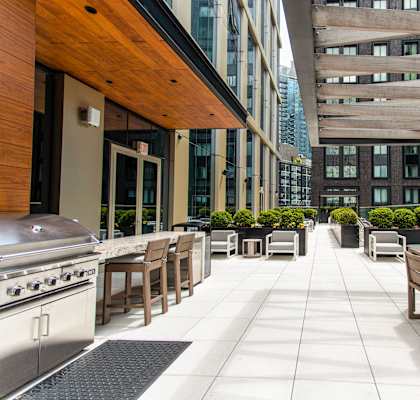 A patio with wooden furniture and a grill is surrounded by buildings.