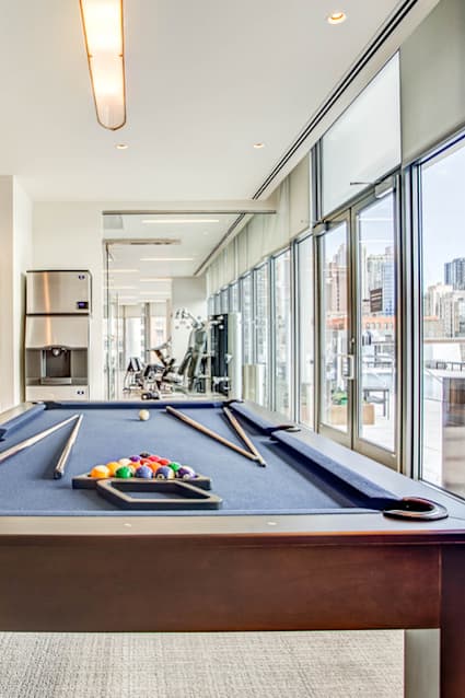 A pool table with balls and cues is in the foreground of a room with a television, couch, and a view of the city.