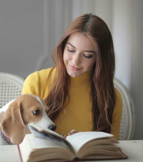 A woman in a yellow sweater is reading a book with a dog sitting beside her.