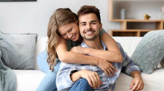 A man and a woman are sitting on a couch, with the woman hugging the man from behind.