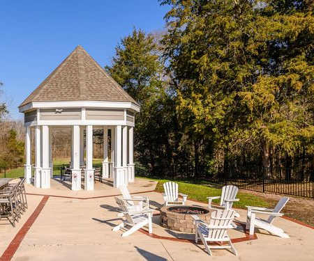The pavilion with a grilling station, a gazebo with tables and chairs, a bar counter with barstool chairs, and a firepit surrounded by white chairs, all encompassed by a grassy area with trees at Retreat at McAlpine Creek Apartment Homes.   