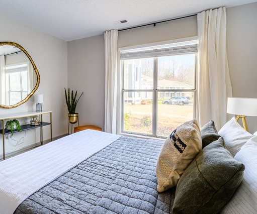 A spacious bedroom with a queen-size bed, a dresser, decorations, large window, and nightstands at Retreat at McAlpine Creek Apartment Homes. 
