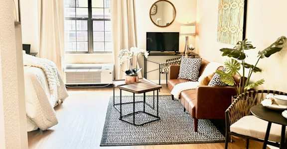 A living room with a brown sofa, a white bed, and a TV on the wall.