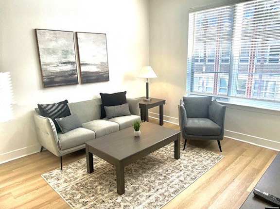 A living room with a grey couch, a black and white painting, a grey armchair, a small table with a lamp and a plant on it, and a large window with blinds.