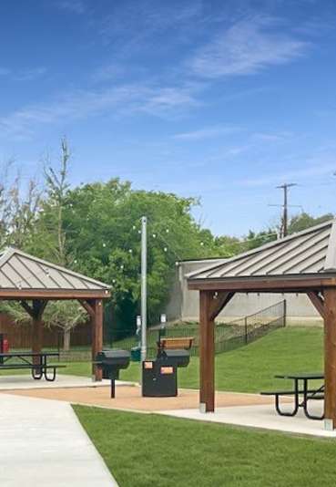 Two wooden gazebos with picnic tables are situated in a grassy park.