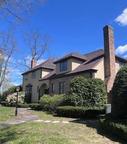 A house with a brown roof and a chimney is surrounded by green bushes and trees.