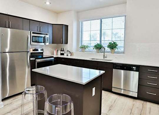Townhomes Near Downtown Phoenix, AZ - The Highland Apartments - Modern Kitchen With White Quartz Island, Dark Cabinets, Stainless Steel Appliances, and Large Window Over Sink.