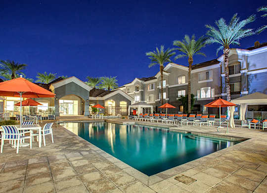 Dog-Friendly Townhomes in Phoenix, AZ - The Highland Apartments - Resort-Style Pool At Night With Palm Trees, Lounge Chairs, Umbrellas, and Apartment Buildings Surrounding The Courtyard.