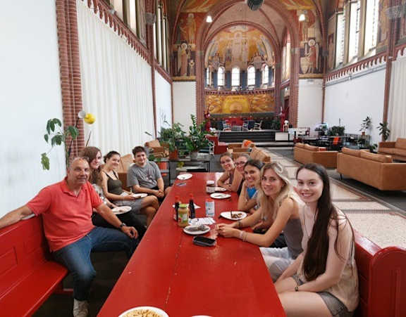A group of people are sitting at a long red table in a large room with a high ceiling and arched windows.
