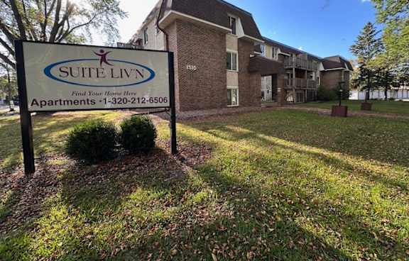 A sign for Suite Livin apartments stands in front of a brick building.