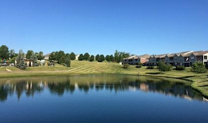 A serene lake reflects the sky and the surrounding greenery and buildings.