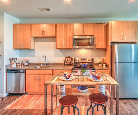 A large, modern kitchen with stainless steel appliances, kitchen utensils, light countertops with lots of room, a kitchen island with barstools and two places set for a meal, and large medium wooden cabinets at Stitchweld Apartment Homes. 
