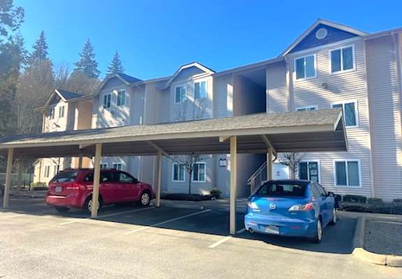 A red car is parked in a covered parking lot in front of a two-story apartment building.