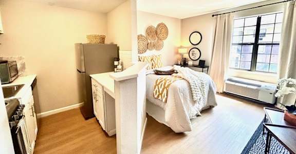 A kitchen with white appliances and a small bedroom area with a bed and a nightstand.