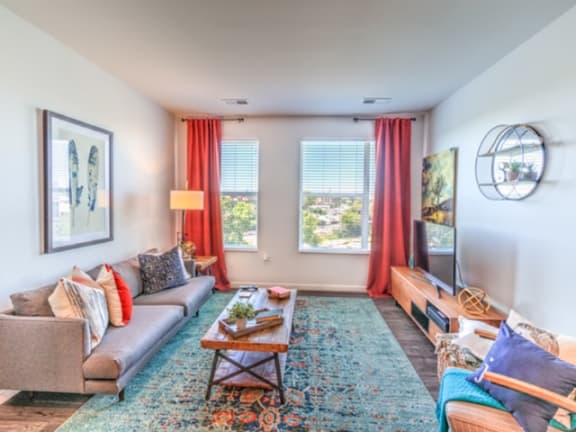 The spacious living room with a gray couch, throw pillows, large windows, a ceiling fan, wall art, plush chair, coffee table, decorations, large windows with red curtains, a TV, and a turquoise rug at Stitchweld Apartment Homes. 