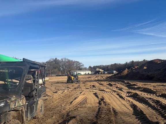 A black ATV is parked on a dirt road with a yellow tractor in the background.