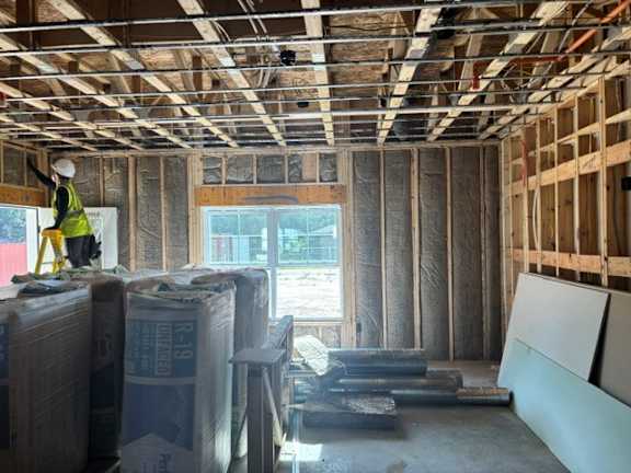 A construction worker in a yellow vest is standing in a room under construction with exposed wooden beams and insulation panels on the walls.