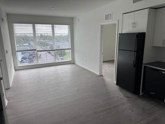 A black fridge sits in a kitchen with a large window overlooking a parking lot.