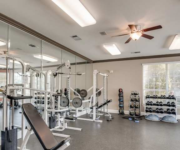 A spacious gym with cardio machines, overhead lighting with a ceiling fan, windows, TVs, and weights at Sycamore Farms Apartment Homes. 