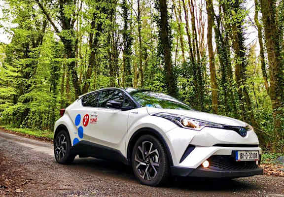 A white car with blue dots and the letters "KO" on it is parked on a dirt road in a forest.