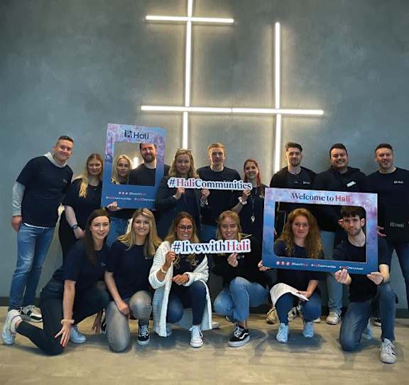 A group of people are gathered in front of a wall with a cross, holding signs that say "#HaliCommunities" and "#livewithHali".
