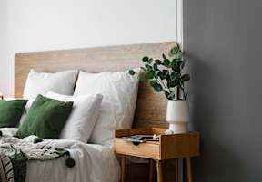 A bed with white pillows and a green throw is next to a wooden nightstand with a plant on it.