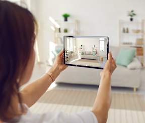 A woman is holding a tablet with a virtual room displayed on the screen.