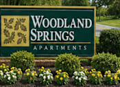 Thumbnail 23 of 24 - A sign for Woodland Springs Apartments is surrounded by flowers.