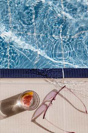 A straw hat, sunglasses, and a sunscreen bottle are placed on a towel by a pool.