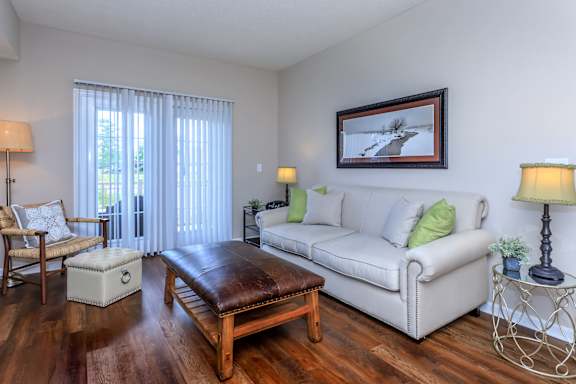 A living room with a white couch, a brown leather ottoman, and a painting on the wall.