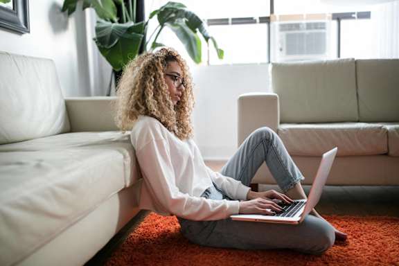 A woman with curly hair is sitting on the floor using a laptop.