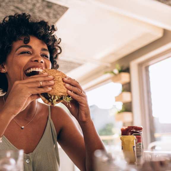 A woman with curly hair is laughing while eating a sandwich.