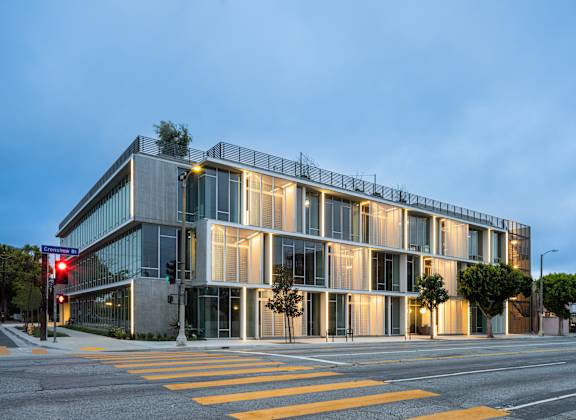 A modern building with a glass facade is illuminated from within and surrounded by a yellow crosswalk.