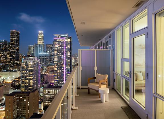 A balcony with a chair and a table overlooking a city skyline at night. at Olympic + Hill Apartments, Apartments, Los Angeles, CA