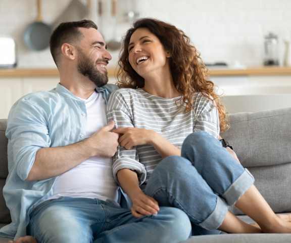 A man and a woman are sitting on a couch, holding hands and smiling at each other.