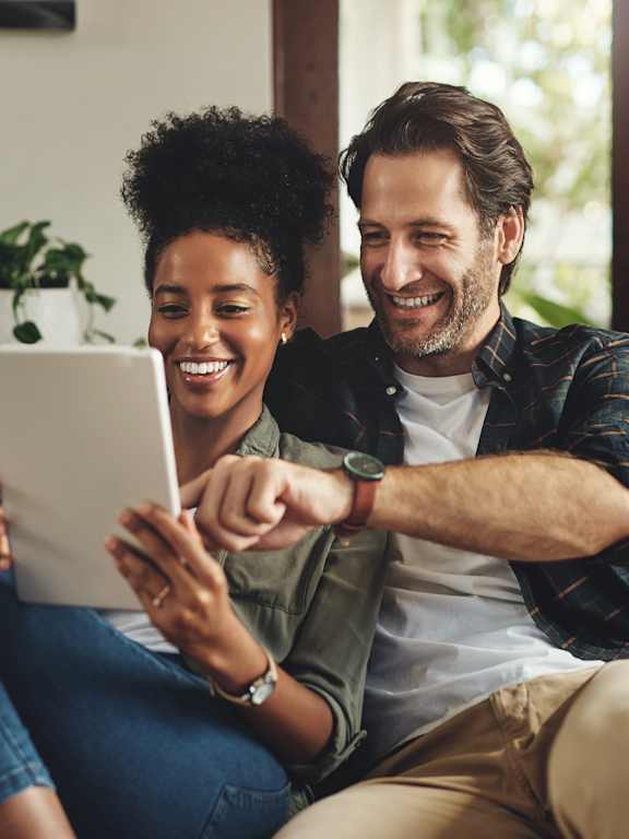 A man and a woman are sitting on a couch, looking at a tablet together.