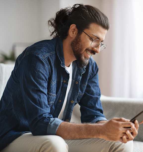 A man with a beard and glasses is sitting on a couch, looking at his phone.