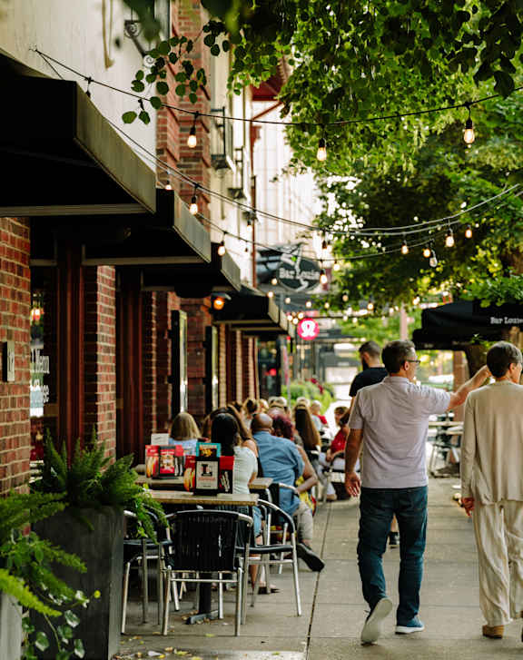 Two men walking down a street with tables and chairs on the side.
