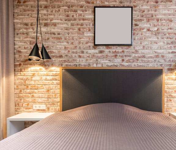 A bed with a pink cover is in front of a brick wall with a white frame hanging on it.