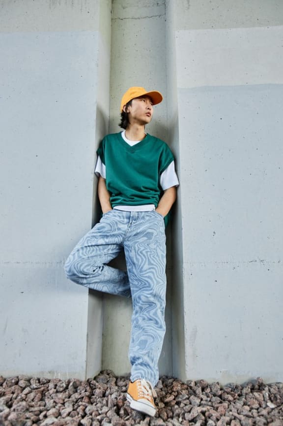 a man leaning against a wall wearing a green shirt and jeans and a yellow hat