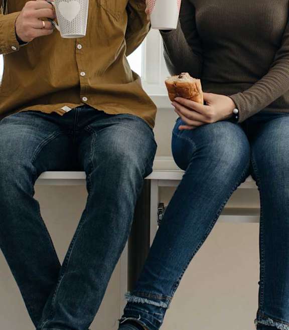 Two people sitting on stools, one holding a mug and the other holding a sandwich.