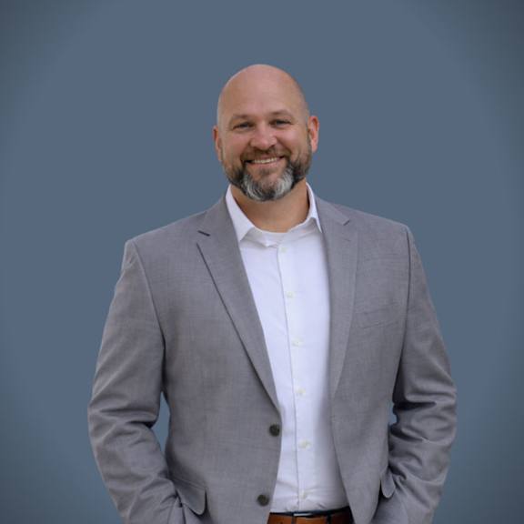 A man with a beard is wearing a grey suit and a white shirt.